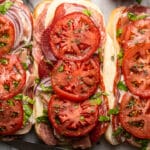 Italian hoagies wit long rolls filled with deli meats, cheese, and veggies topped with olive oil on marble table.