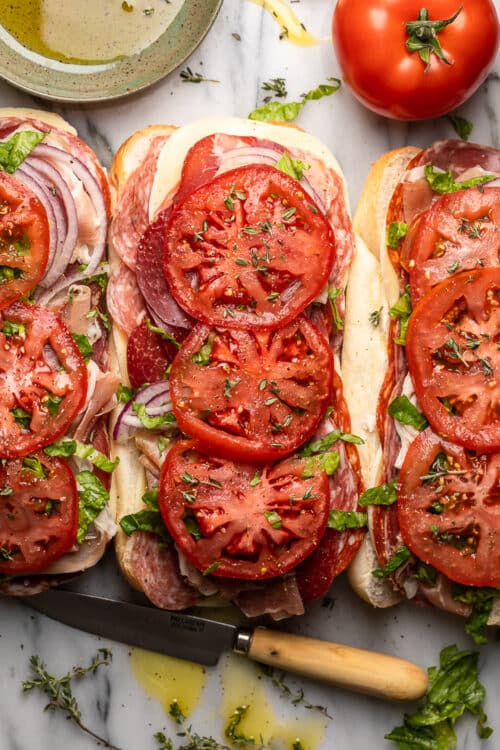 Italian hoagies wit long rolls filled with deli meats, cheese, and veggies topped with olive oil on marble table.