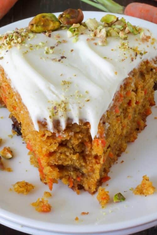 A slice of pumpkin carrot cake with a bite taken out of it. Cream cheese frosting on top with pistachios.