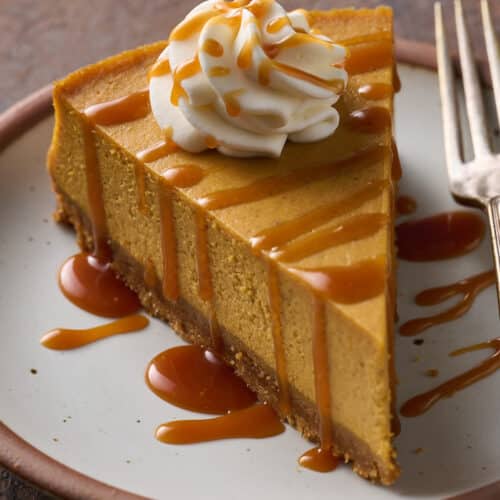 A slice of pumpkin cheesecake on a plate topped with whipped cream and salted caramel sauce.