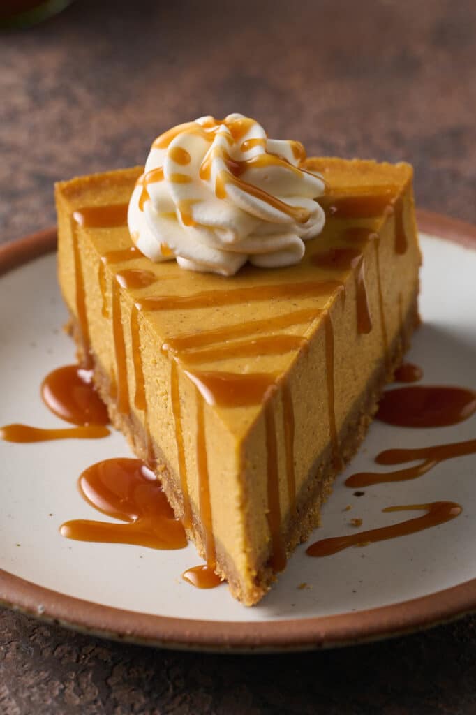 A slice of pumpkin cheesecake on a plate topped with whipped cream and salted caramel sauce.