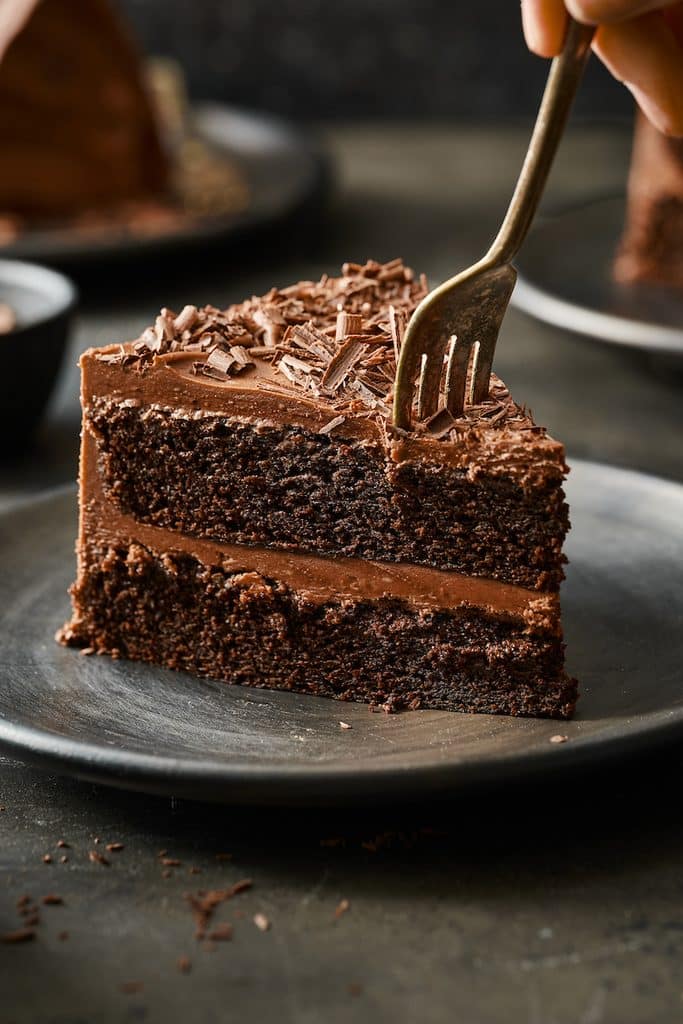 Fork cutting into a slice of chocolate cake topped with chocolate frosting and chocolate shavings.