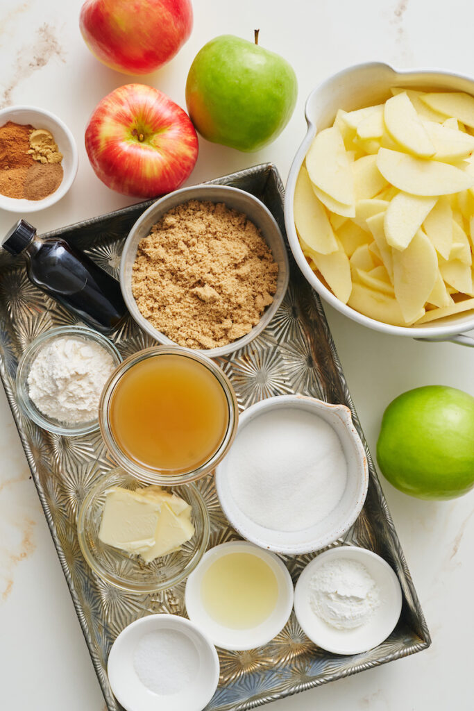 Apple pie ingredients including sliced apples, brown sugar, and apple cider, on a baking sheet. 