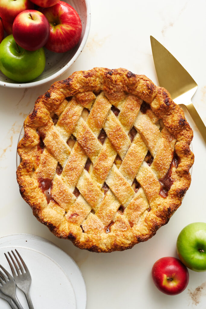 Whole apple pie recipe on a table with apples and a pie sever. 