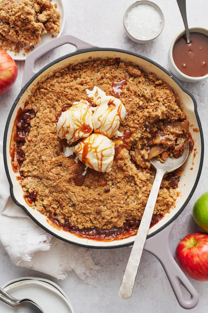 Apple crisp recipe in a large skillet topped with ice cream and a drizzle of caramel sauce.