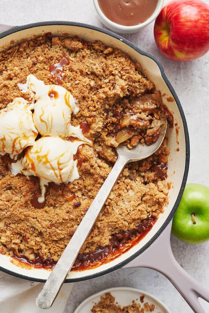 Apple crisp recipe in a large skillet topped with ice cream and a drizzle of caramel sauce.