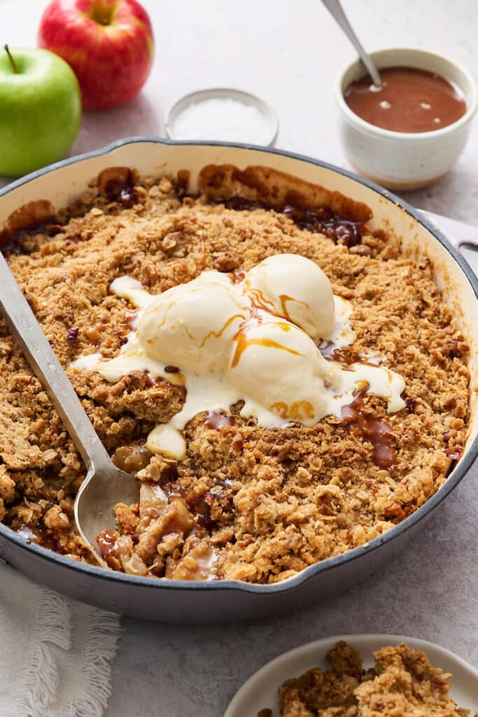 A large baking dish filled with apple crisp topped with a scoop of vanilla ice cream and a drizzle of caramel sauce. 