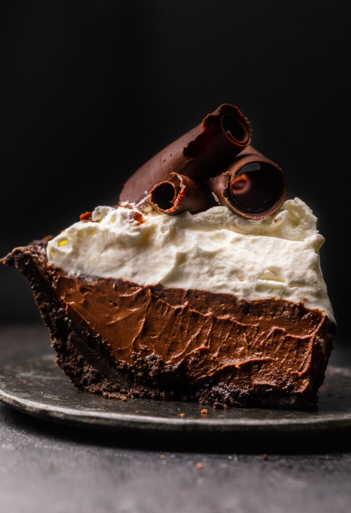 Slice of chocolate pudding pie with whipped cream topping and chocolate curls on a plate.