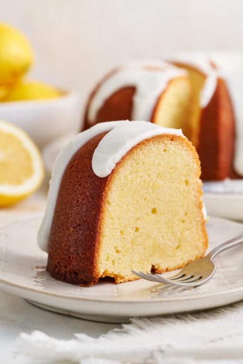 Slice of lemon bundt cake on white plate with fork and lemon slices.