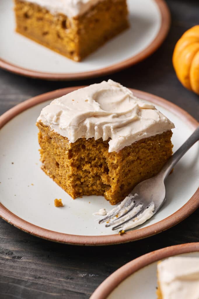 A slice of pumpkin cake on a white plate with a bite taken out of it.