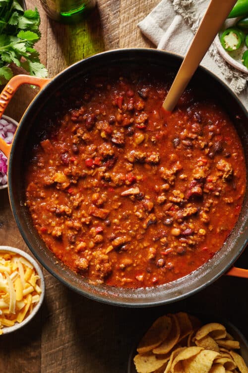 A dutch oven with homemade chili in it. Chili toppings on the table below.