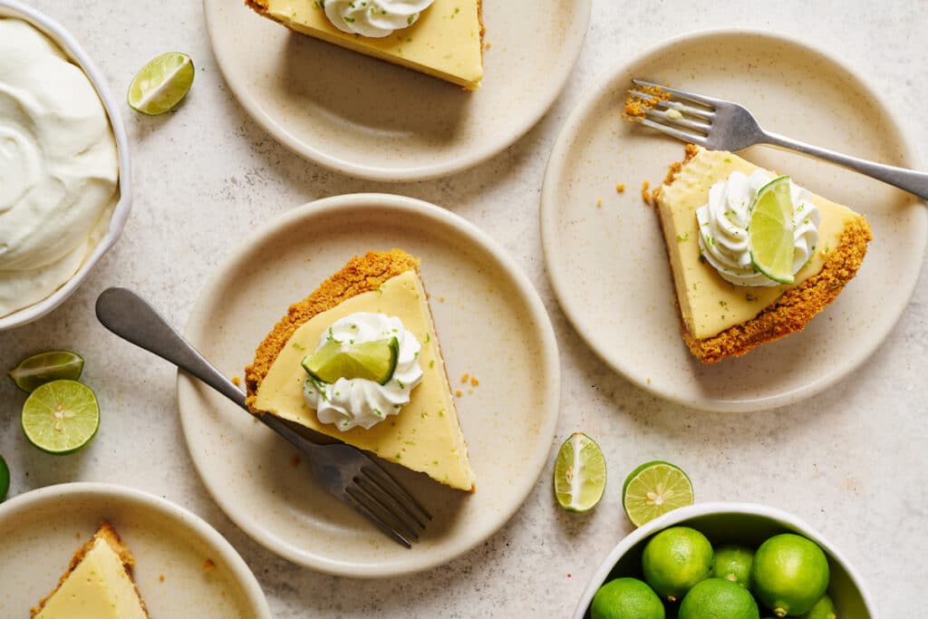 Slices of key lime pie on a table with limes and whipped cream on the side.