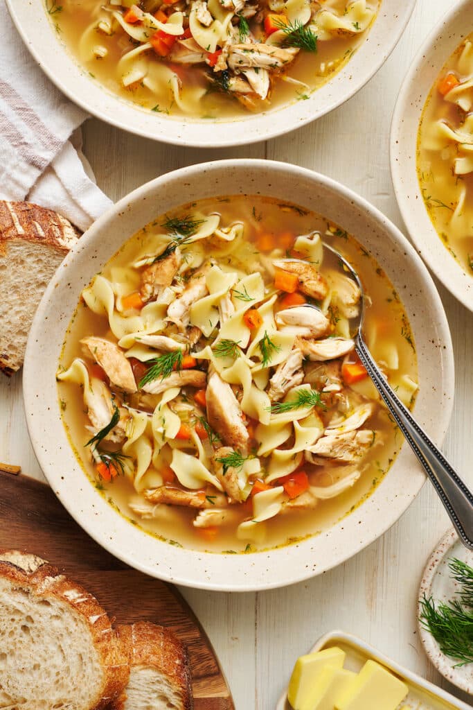 Chicken noodle soup in a white bowl with a soup spoon and bread on the side.