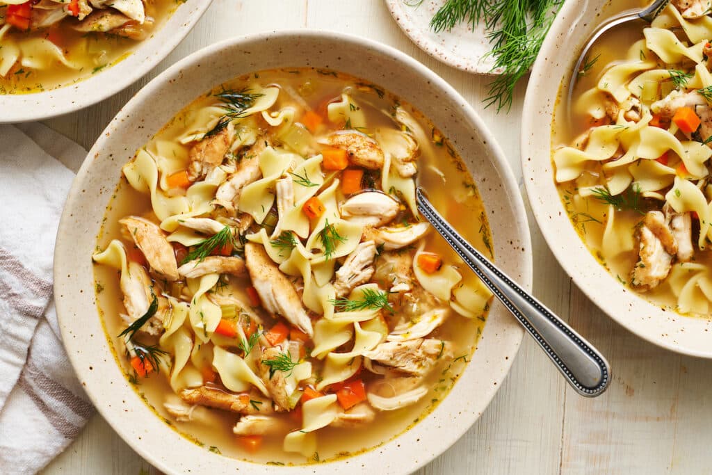 Chicken noodle soup in a white bowl with a soup spoon and bread on the side.