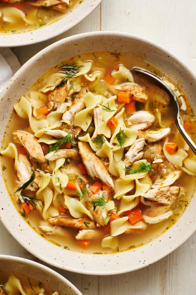 Chicken noodle soup in a white bowl with a soup spoon and bread on the side.