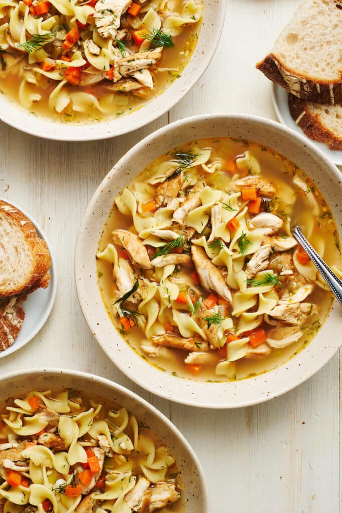 Chicken noodle soup in a white bowl with a soup spoon and bread on the side.