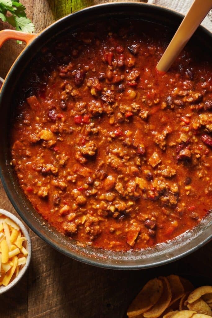 A dutch oven with ground beef homemade chili in it. Chili toppings on the table below.
