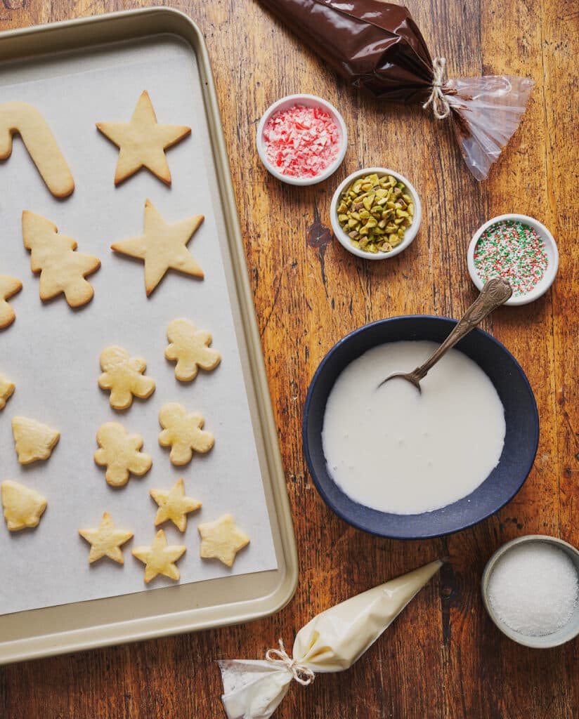 Baked Christmas cookies on baking sheet with icing and decorations on the side.