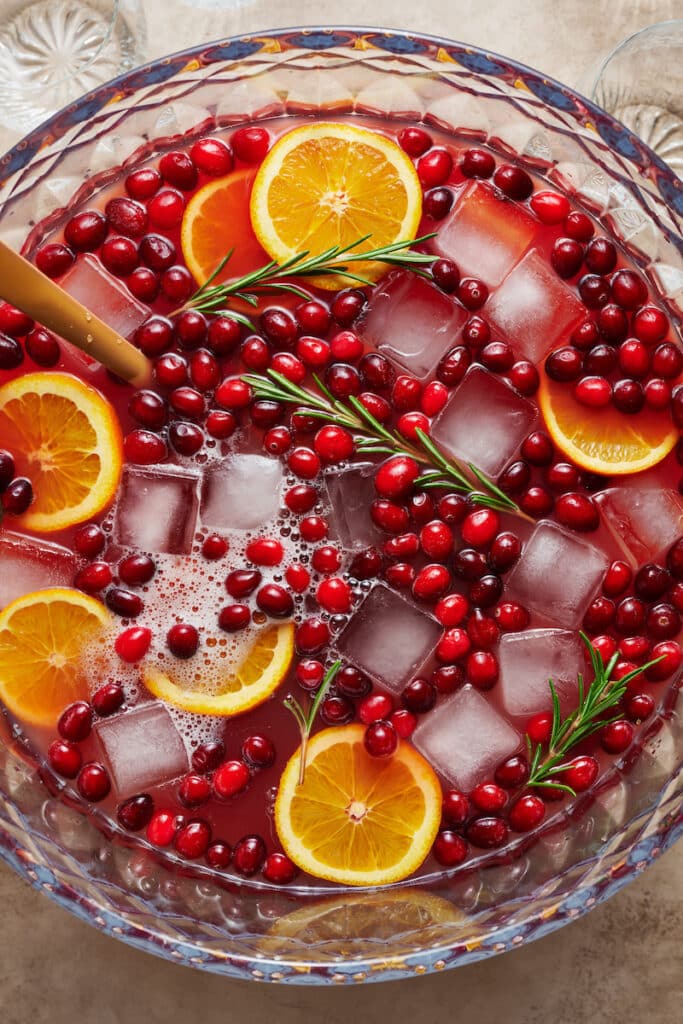 A punch bowl full of Christmas punch topped with fresh cranberries, orange slices, and rosemary.