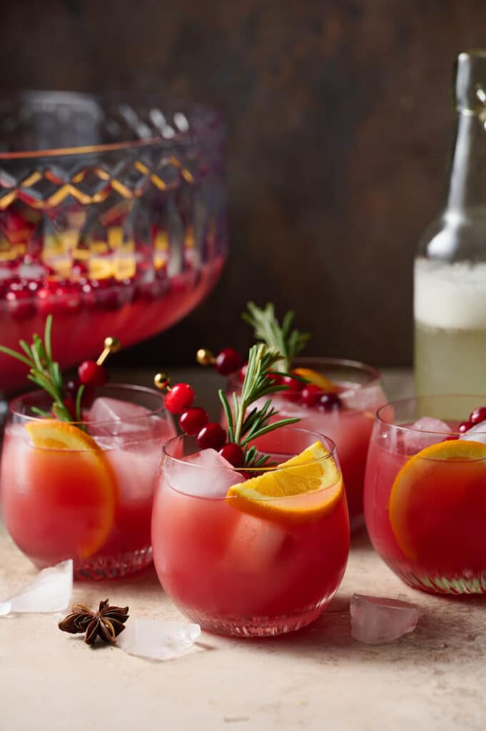 Glasses full of Christmas punch garnished with sprigs of rosemary, slices of orange, and cranberries.