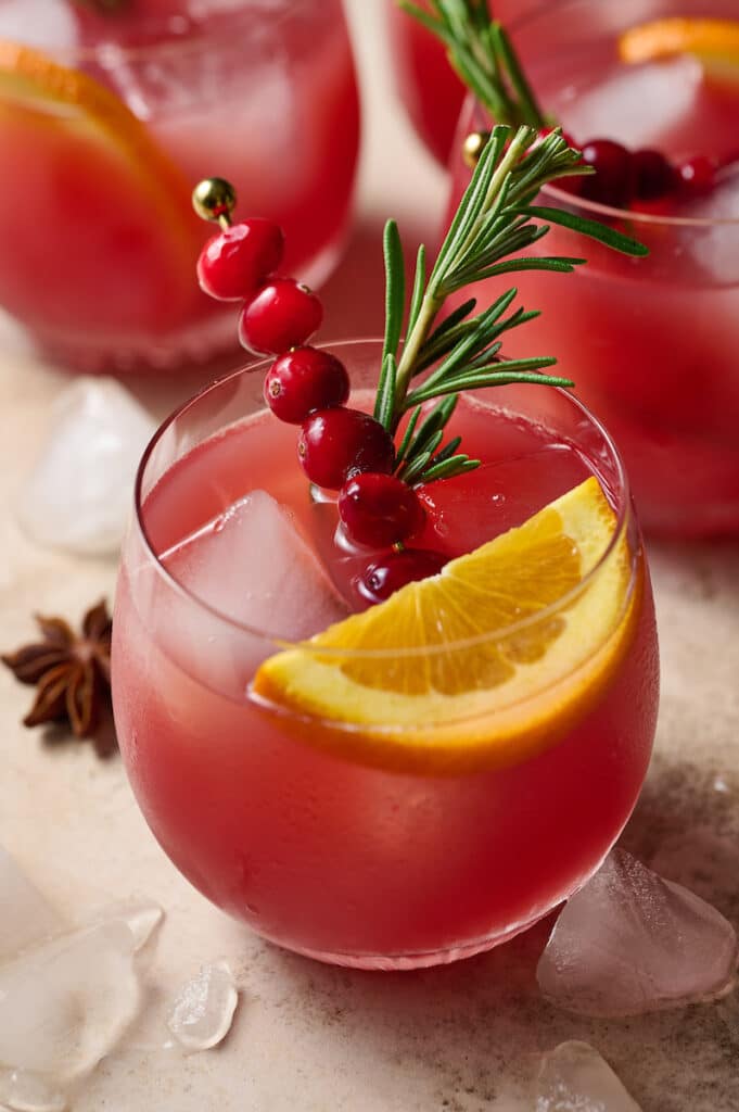 Glasses full of Christmas punch garnished with sprigs of rosemary, slices of orange, and cranberries.