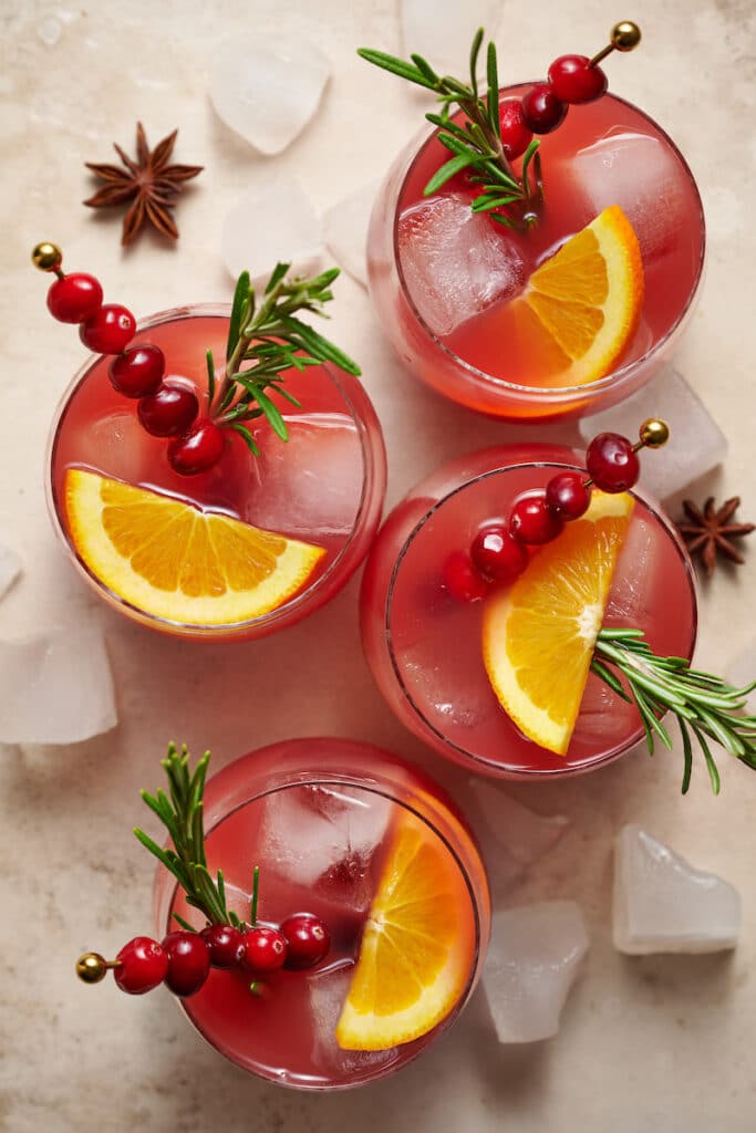 Glasses full of Christmas punch garnished with sprigs of rosemary, slices of orange, and cranberries.