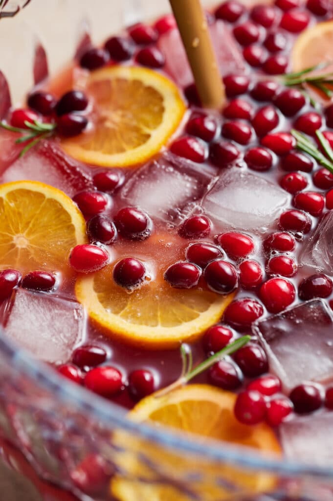 A punch bowl full of Christmas punch topped with fresh cranberries, orange slices, and rosemary.