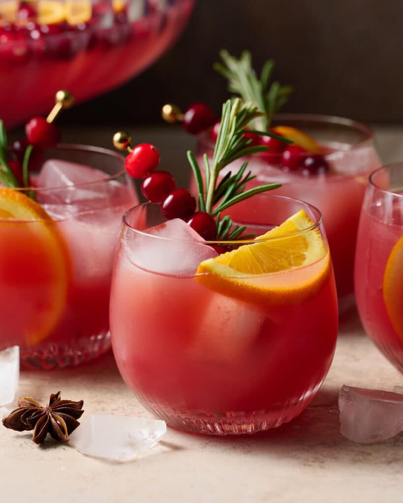 Glasses full of Christmas punch garnished with sprigs of rosemary, slices of orange, and cranberries.