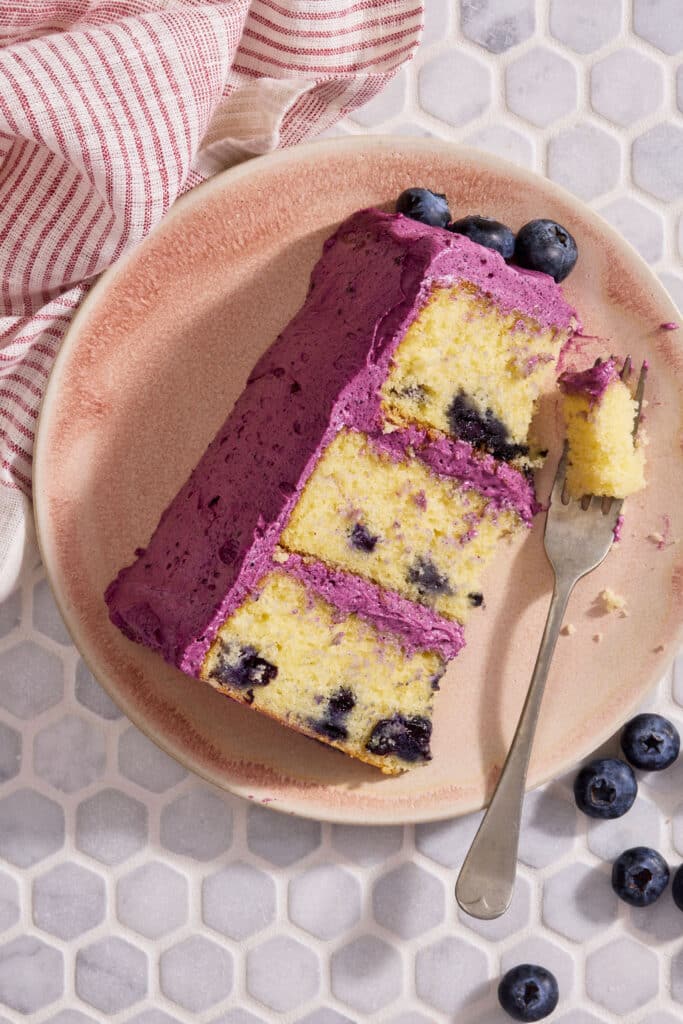 A slice of lemon blueberry cake covered with blueberry frosting and topped with fresh blueberries.